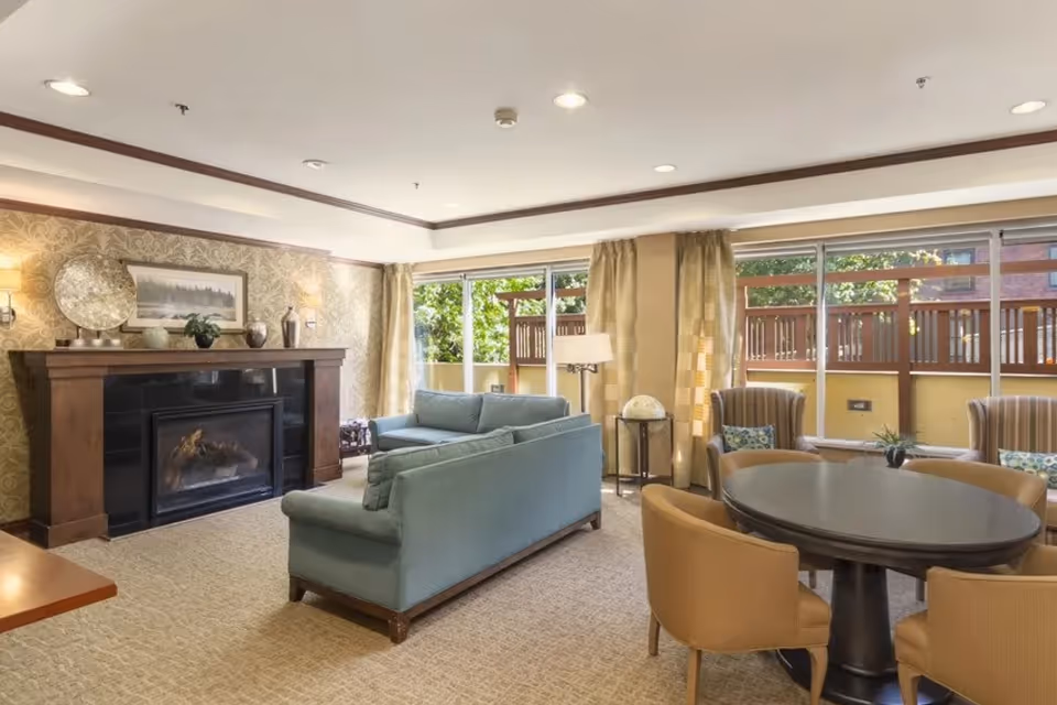 Bright common living room with a fireplace, blue sofas, a round table with chairs, and large windows overlooking an outdoor patio.