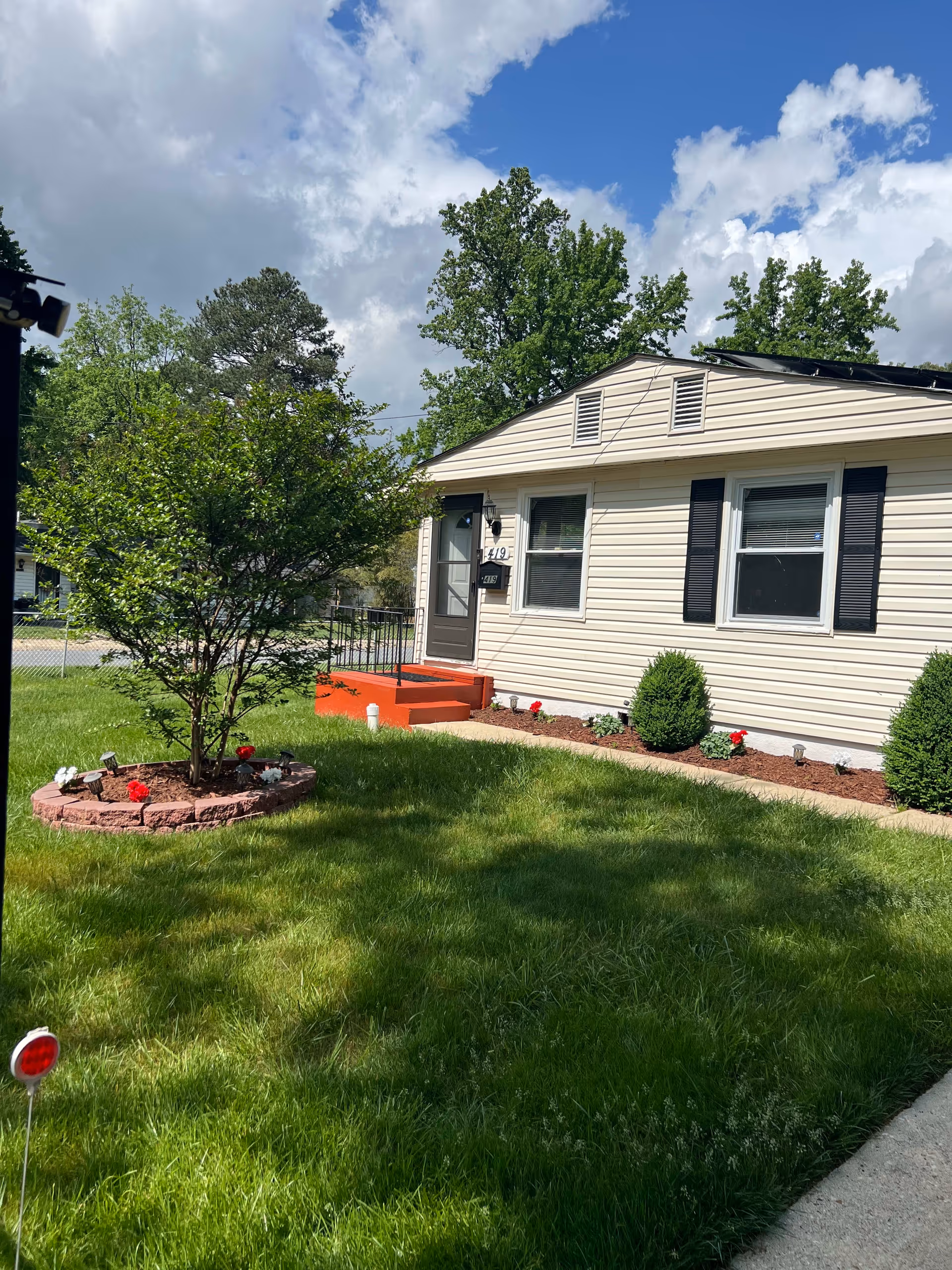 Exterior view of a single-story beige house with black shutters and a small porch with red steps. The house is surrounded by a well-maintained green lawn, a small tree planted in a circular brick planter, and some bushes and flowers along the foundation. The sky is partly cloudy with patches of blue.