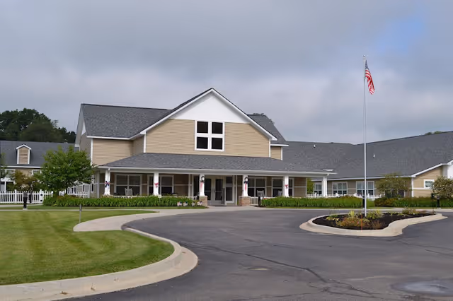 Front exterior of a beige senior living facility with a covered entrance, circular driveway, landscaped lawn, and an American flag.