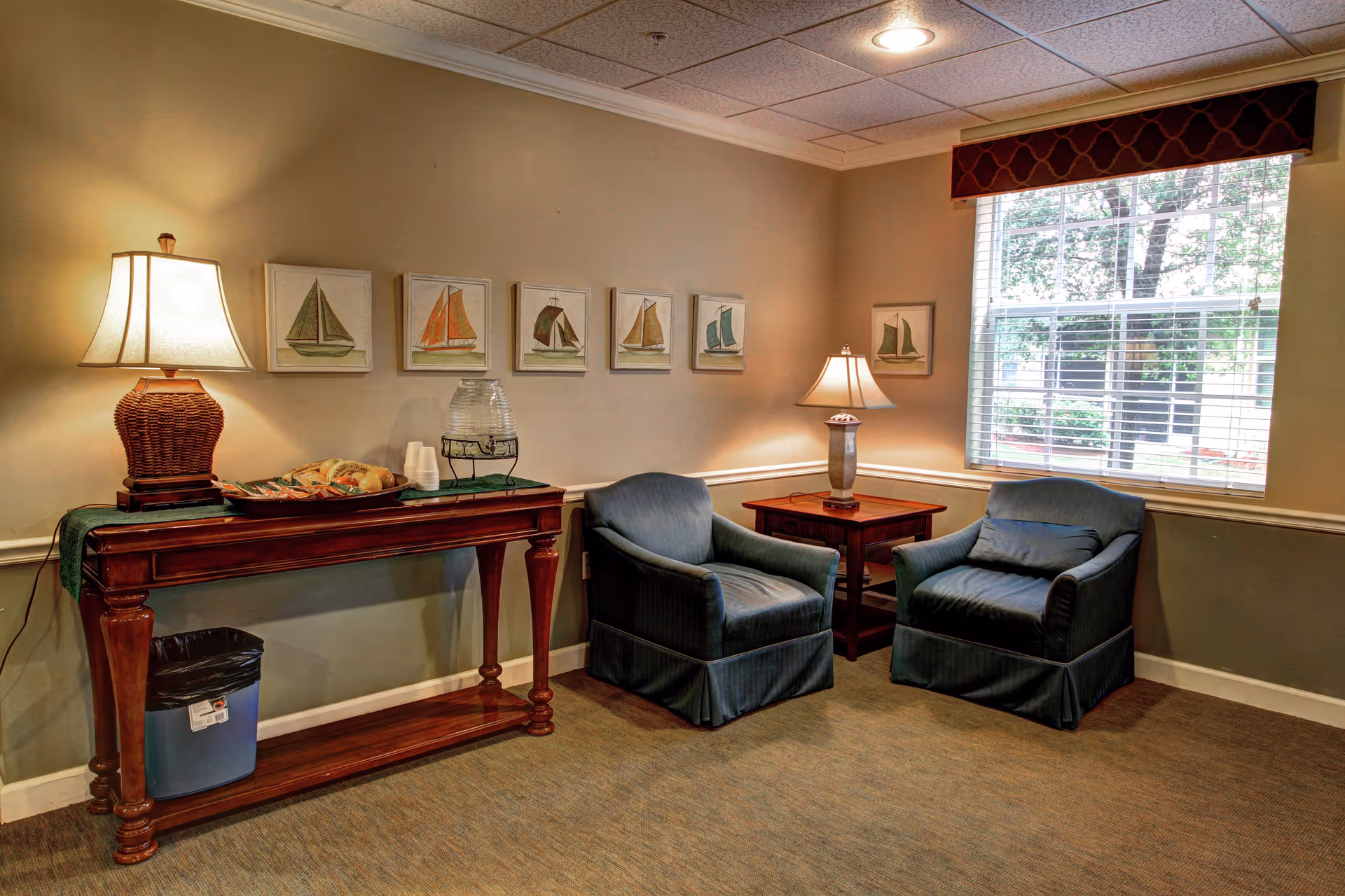 A cozy sitting area with two blue upholstered armchairs and a wooden side table with a lamp between them. A large window with blinds and a valance lets in natural light. On the opposite wall, there is a wooden console table with a lamp, a tray of bread rolls, a water dispenser, and some cups. Above the console table, five framed pictures of sailboats are hung on the wall. The room has beige walls and carpeted floor.