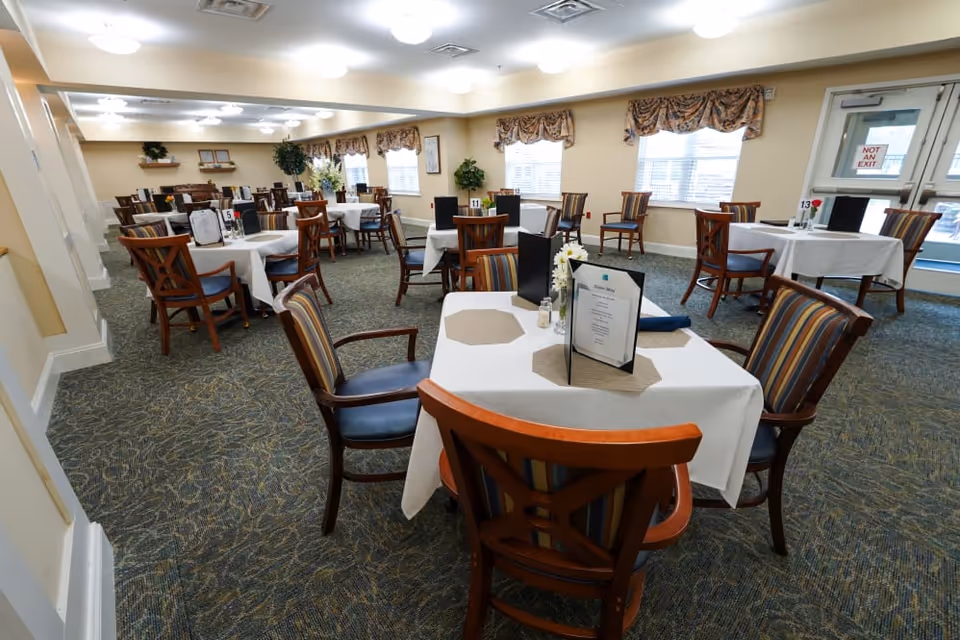 A spacious dining room with multiple tables covered in white tablecloths and set with menus and small flower arrangements. The room has carpeted floors, beige walls, and several windows with patterned valances allowing natural light to enter. Wooden chairs with striped cushions surround each table, and ceiling lights illuminate the space evenly.