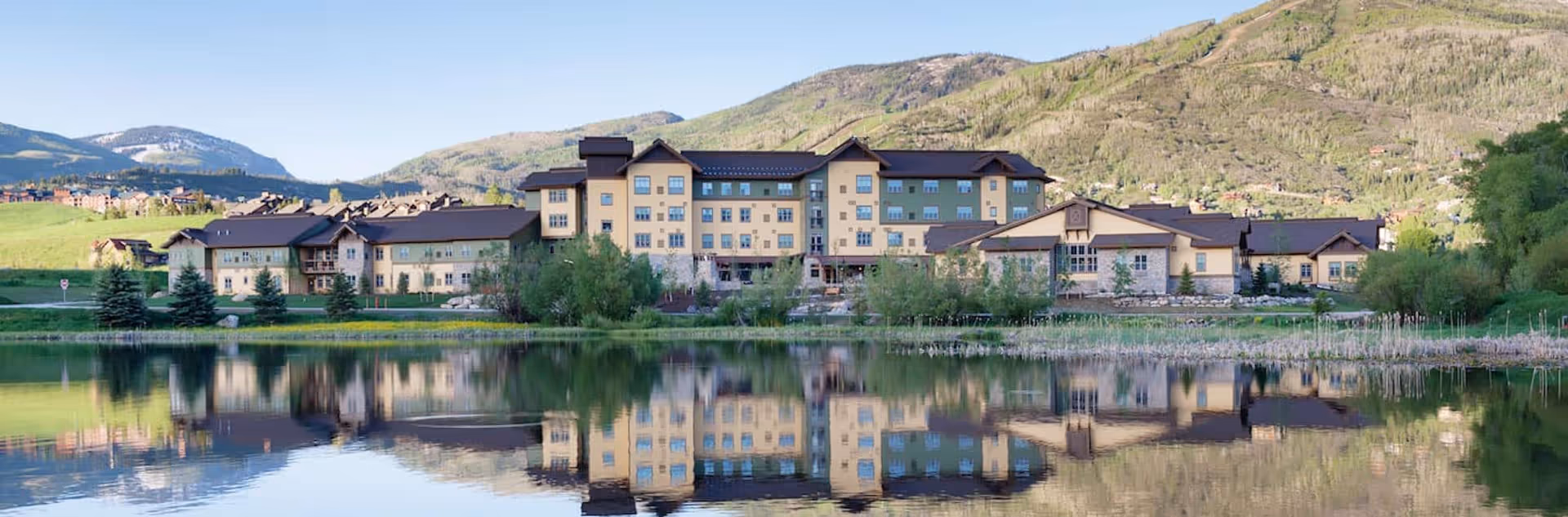A large multi-story building with beige and green exterior walls and dark roofs, situated in a scenic mountainous area. The building is reflected in a calm body of water in the foreground, surrounded by trees and greenery under a clear blue sky.