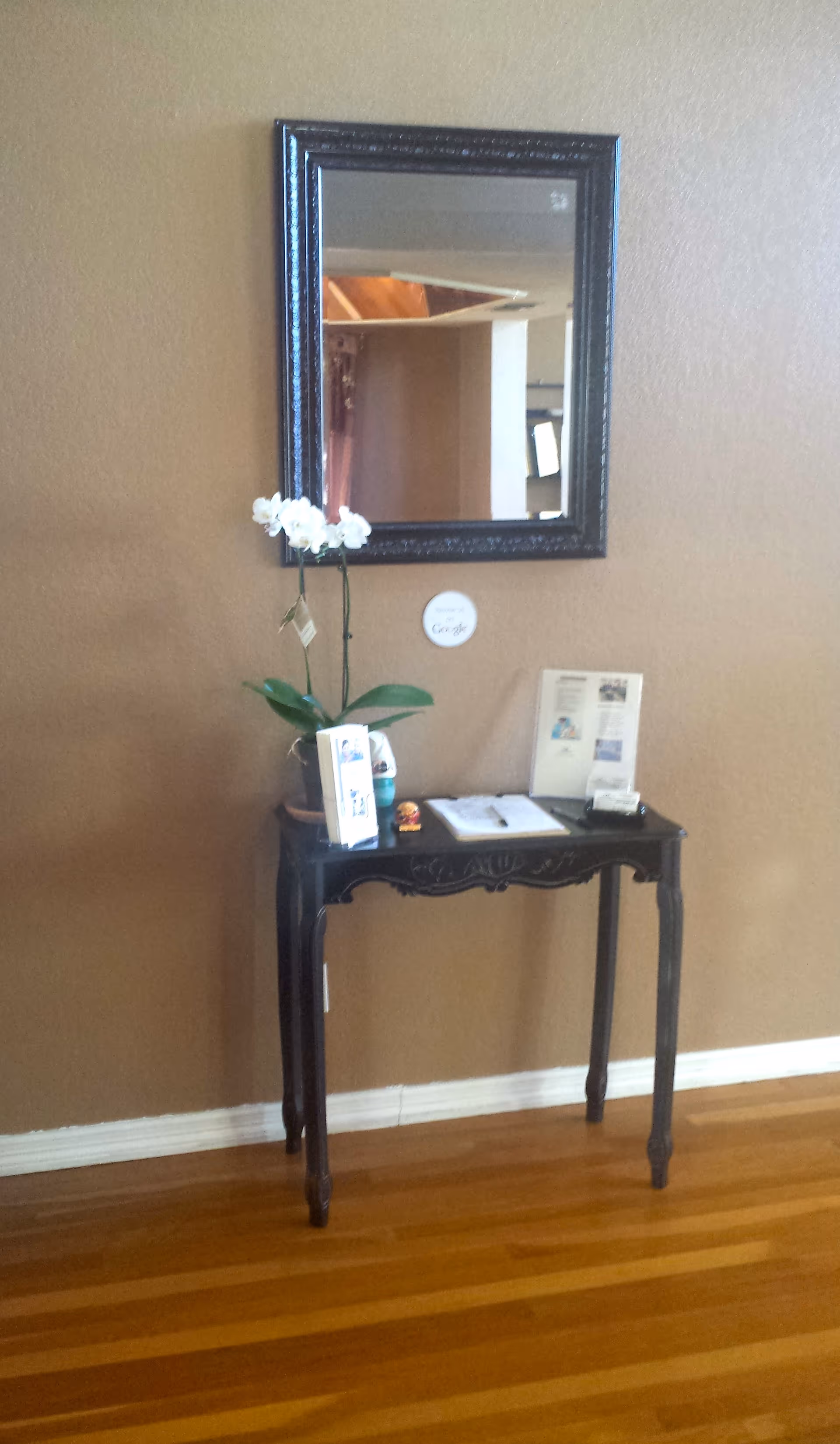 A small black wooden table with carved details stands against a beige wall on a hardwood floor. On the table are a potted white orchid, a few brochures or pamphlets, a small figurine, a pen on an open guestbook, and some other small items. Above the table hangs a rectangular mirror with a dark ornate frame reflecting part of the room.