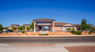 Front exterior view of Advanced Health Care of Albuquerque building with a clear blue sky, landscaped area with bushes and trees, and a paved road in the foreground.