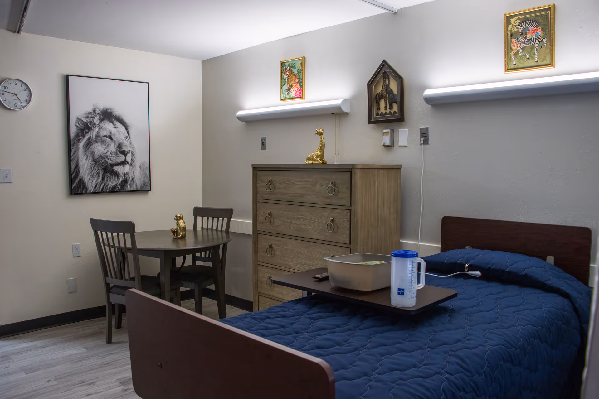 A senior living facility bedroom with a single bed covered in a dark blue quilt. There is a wooden dresser with a golden giraffe figurine on top, a small round table with two chairs, and a golden monkey figurine on the table. The walls are decorated with framed animal artwork including a lion, a zebra, and a giraffe. The room has light-colored walls and flooring, with overhead lighting above the dresser and bed.
