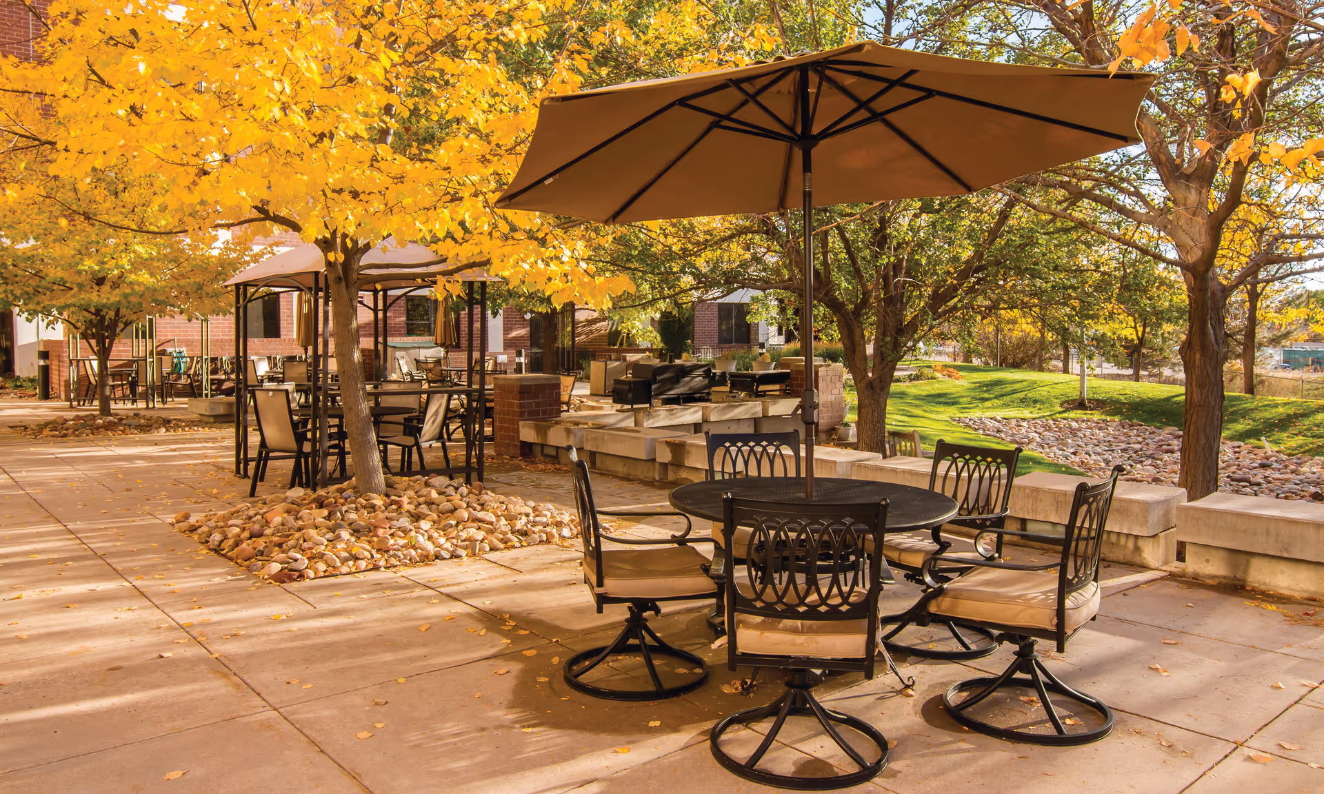 Outdoor patio area with metal table and cushioned chairs under a large umbrella, surrounded by trees with yellow and green leaves, and a building in the background.