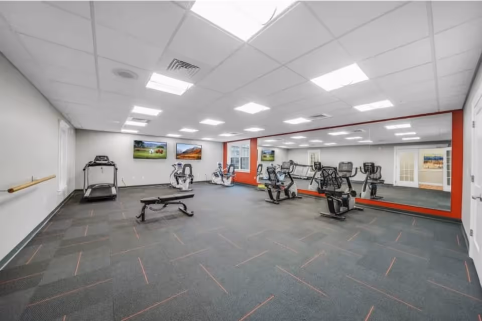 A spacious fitness room with exercise equipment including a treadmill, stationary bikes, and a workout bench. The room has a large mirror on one wall, bright overhead lighting, and two landscape paintings on the far wall.