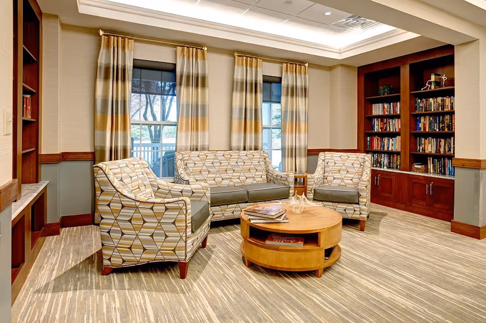 A cozy living room area with a patterned sofa and two matching armchairs arranged around a round wooden coffee table. Behind the seating are three windows with striped curtains. To the right is a built-in wooden bookshelf filled with books and decorative items. The room has a carpeted floor and soft lighting from a recessed ceiling.