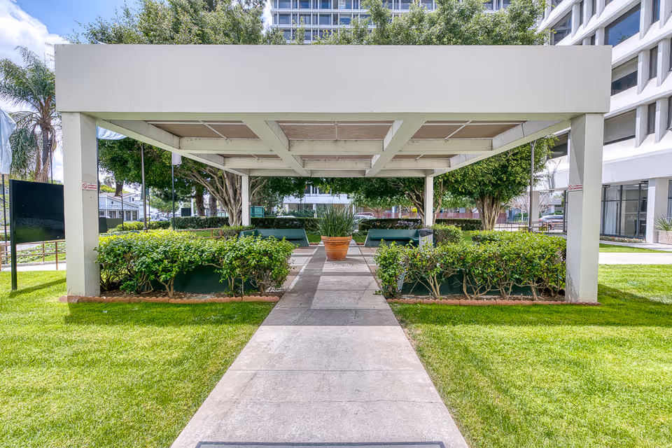 Outdoor covered seating area with benches surrounded by green bushes and trees, located on a grassy lawn with a concrete pathway leading to it. Tall buildings are visible in the background under a partly cloudy sky.