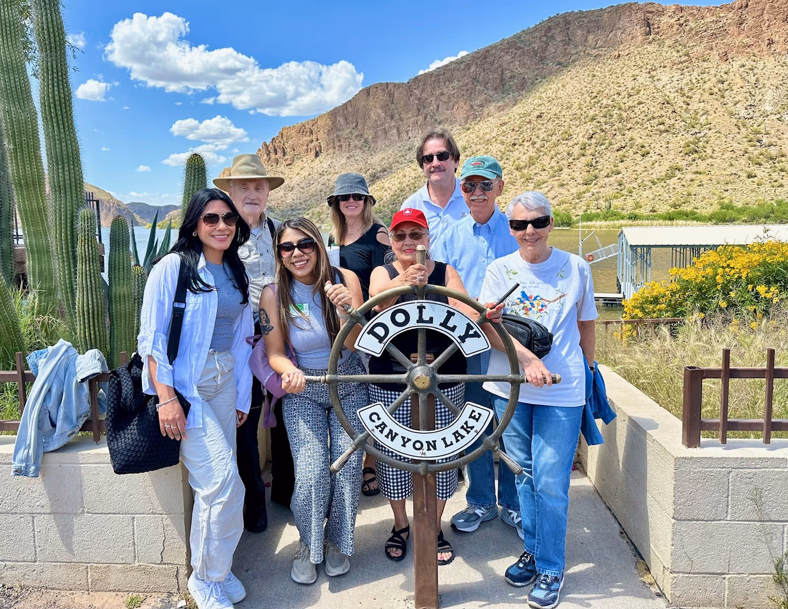 A group of eight people posing outdoors around a large ship's wheel labeled 'DOLLY CANYON LAKE' with a mountainous desert landscape and a lake in the background under a blue sky with scattered clouds.