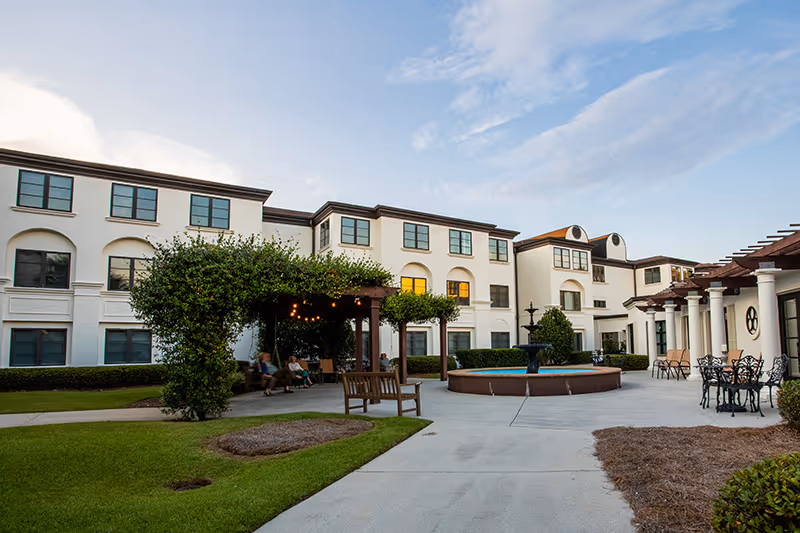 Outdoor courtyard area of a senior living facility with a three-story white building in the background. The courtyard features a central fountain, a pergola with greenery and string lights, benches, and outdoor seating with tables and chairs. The sky is partly cloudy with soft evening light.