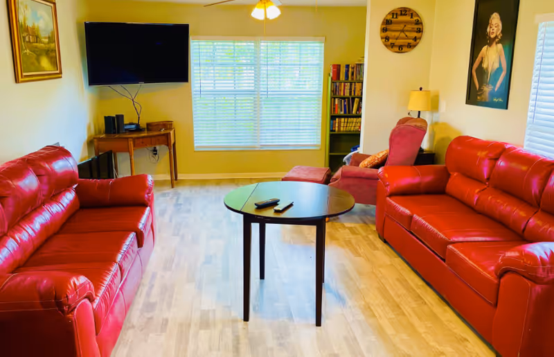 A bright living room with two red leather sofas facing each other, a small round wooden table with two remote controls on it in the center, a wall-mounted flat-screen TV on the left wall, a wooden side table beneath the TV, a large window with blinds letting in natural light, a bookshelf in the corner, a red armchair with an ottoman, a wall clock, a lamp, and a framed picture of a woman on the right wall.