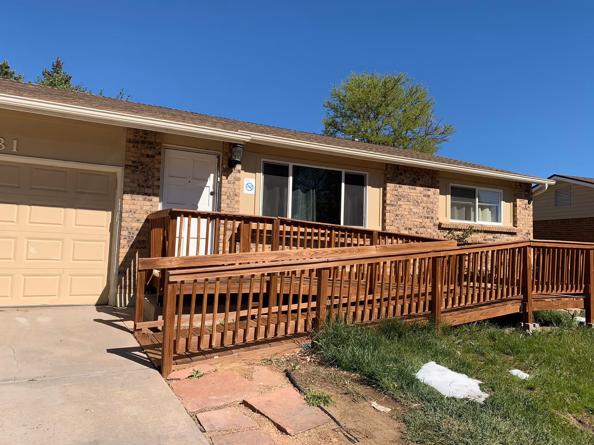 Exterior view of a single-story brick building with a beige garage door and a white entrance door. A wooden wheelchair ramp with railings leads up to the entrance. There is a large window next to the door and a tree in the background under a clear blue sky.
