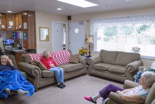 A cozy living room in a senior living facility with three elderly individuals seated on comfortable sofas and armchairs. The room features two brown sofas, a beige armchair, a large window with white lace curtains, a side table with a lamp and a vase of sunflowers, and a small desk area with a computer and cabinets in the background.