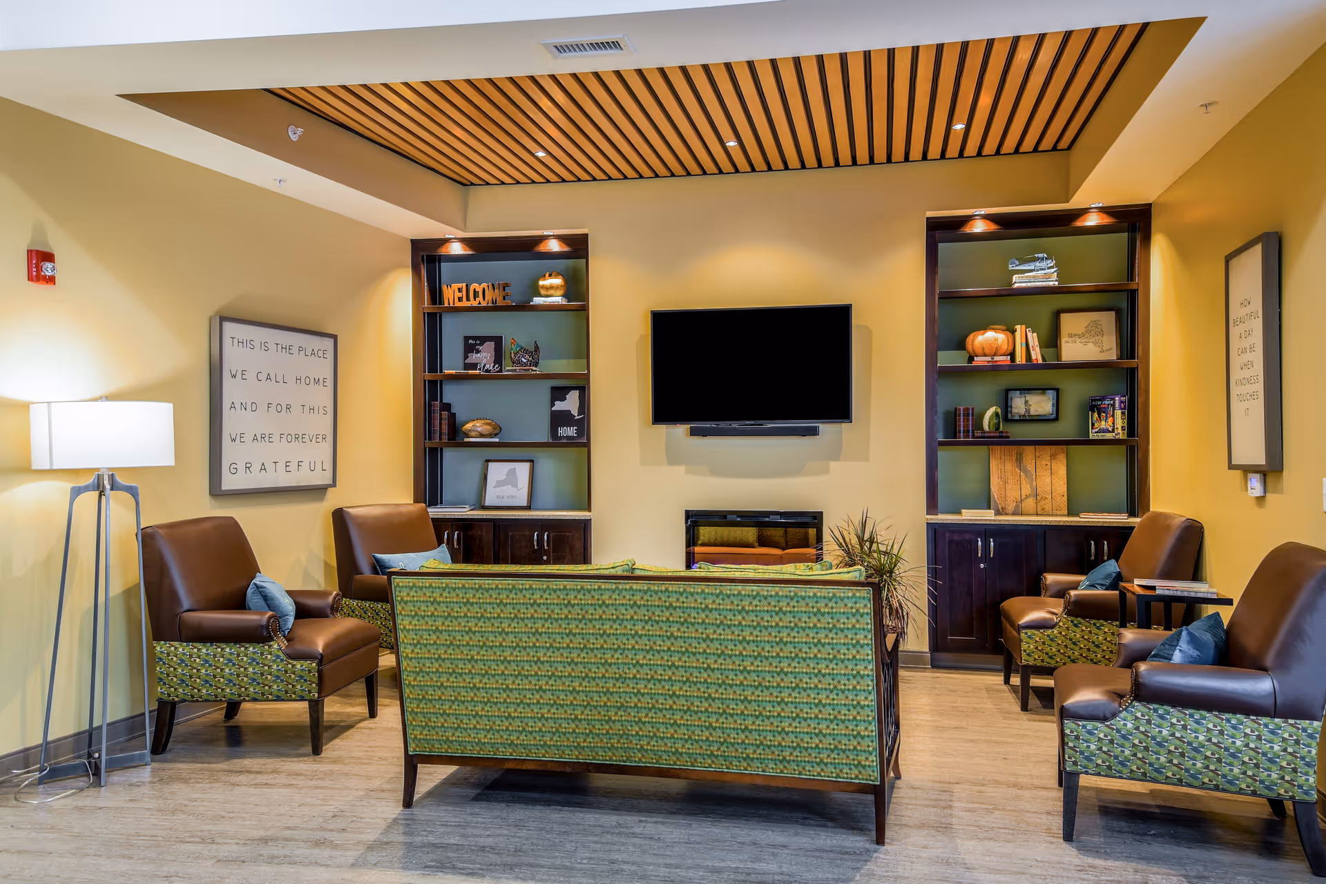 A cozy senior living common area with a green patterned sofa and four brown leather armchairs arranged around a wall-mounted flat screen TV and an electric fireplace. The room features built-in dark wood shelves with decorative items, a floor lamp, and framed inspirational quotes on the yellow walls. The ceiling has a wooden slat design in the center.