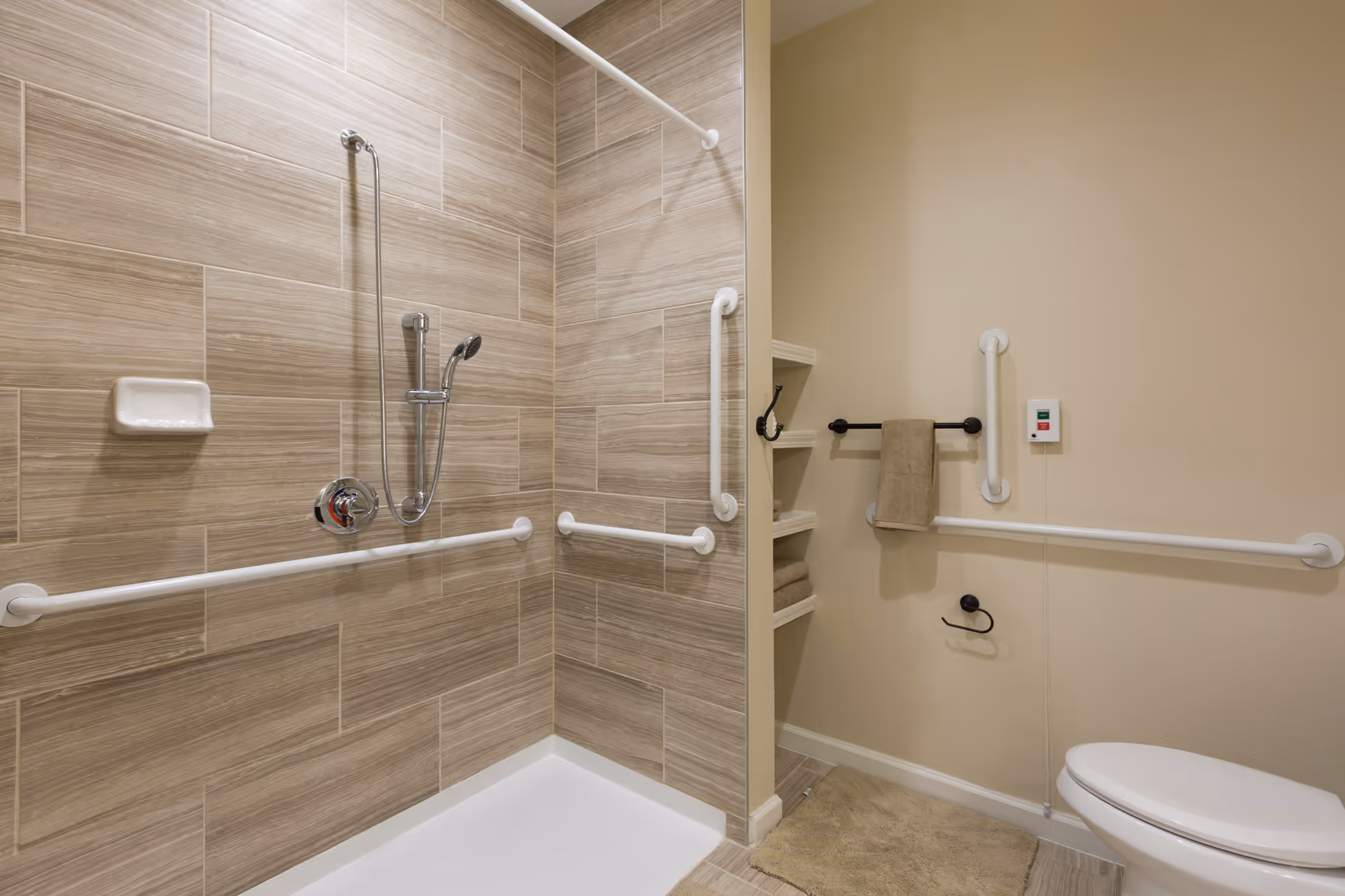 A senior-friendly bathroom featuring a walk-in shower with beige tiled walls, multiple white grab bars for safety, a handheld showerhead, and a soap holder. Adjacent to the shower is a toilet with additional grab bars, a towel rack with a beige towel, open shelving with folded towels, and a beige bath mat on the floor.