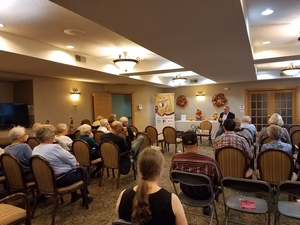 A group of elderly people seated in a room facing a man who appears to be giving a talk or presentation. The room has beige walls, carpeted floor, and ceiling lights. There are decorative wreaths on the wall and a banner with text and social media icons near the speaker.