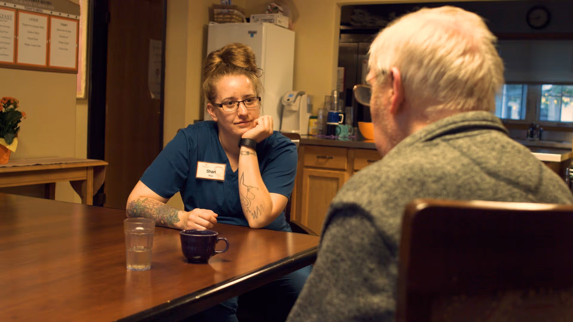 A woman wearing glasses and a blue uniform with a name tag that reads 'Shari' sits at a wooden table, attentively listening to an elderly man with white hair and glasses who is seated across from her. There are a glass of water and a dark-colored mug on the table. The background shows a kitchen area with cabinets, a refrigerator, and various kitchen items.