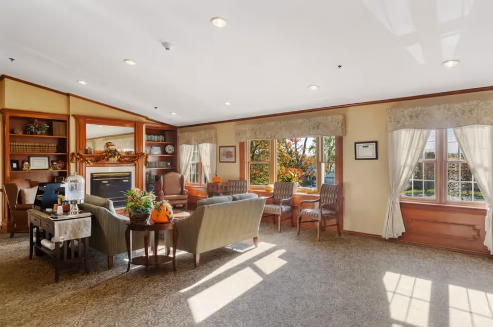 A bright and cozy living room area with large windows letting in natural light. The room features comfortable upholstered chairs and sofas arranged around a fireplace with a wooden mantel decorated with autumn-themed garland. Built-in wooden bookshelves flank the fireplace, holding books and decorative items. The windows have light-colored curtains and valances, and there is a patterned carpet covering the floor.