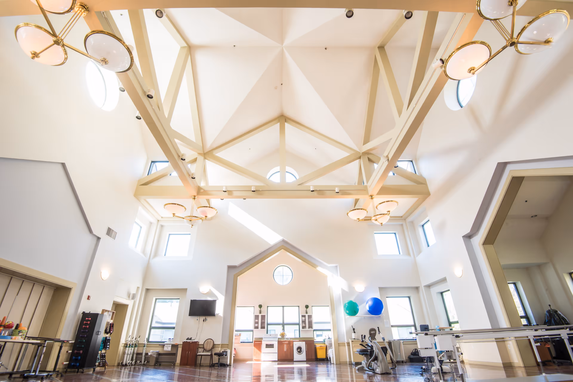 A spacious rehabilitation room with high vaulted ceilings and large windows letting in natural light. The room contains exercise equipment, therapy balls, and a small kitchen area with appliances. The walls are white with beige structural beams and elegant light fixtures hanging from the ceiling.