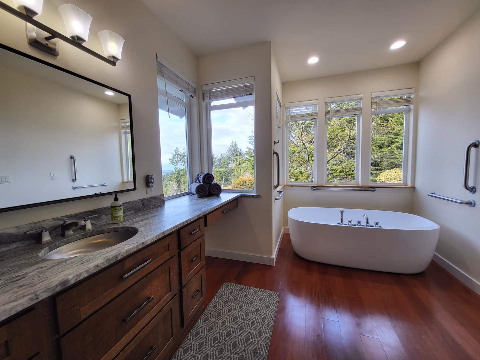 A spacious bathroom with a large wooden vanity featuring a marble countertop and an oval sink. Above the vanity is a large mirror with three modern light fixtures. To the right, there is a white freestanding bathtub positioned near a set of windows that offer a view of green trees outside. The bathroom has wooden flooring and several grab bars installed on the walls for safety. A few rolled towels are placed on the countertop near the windows.