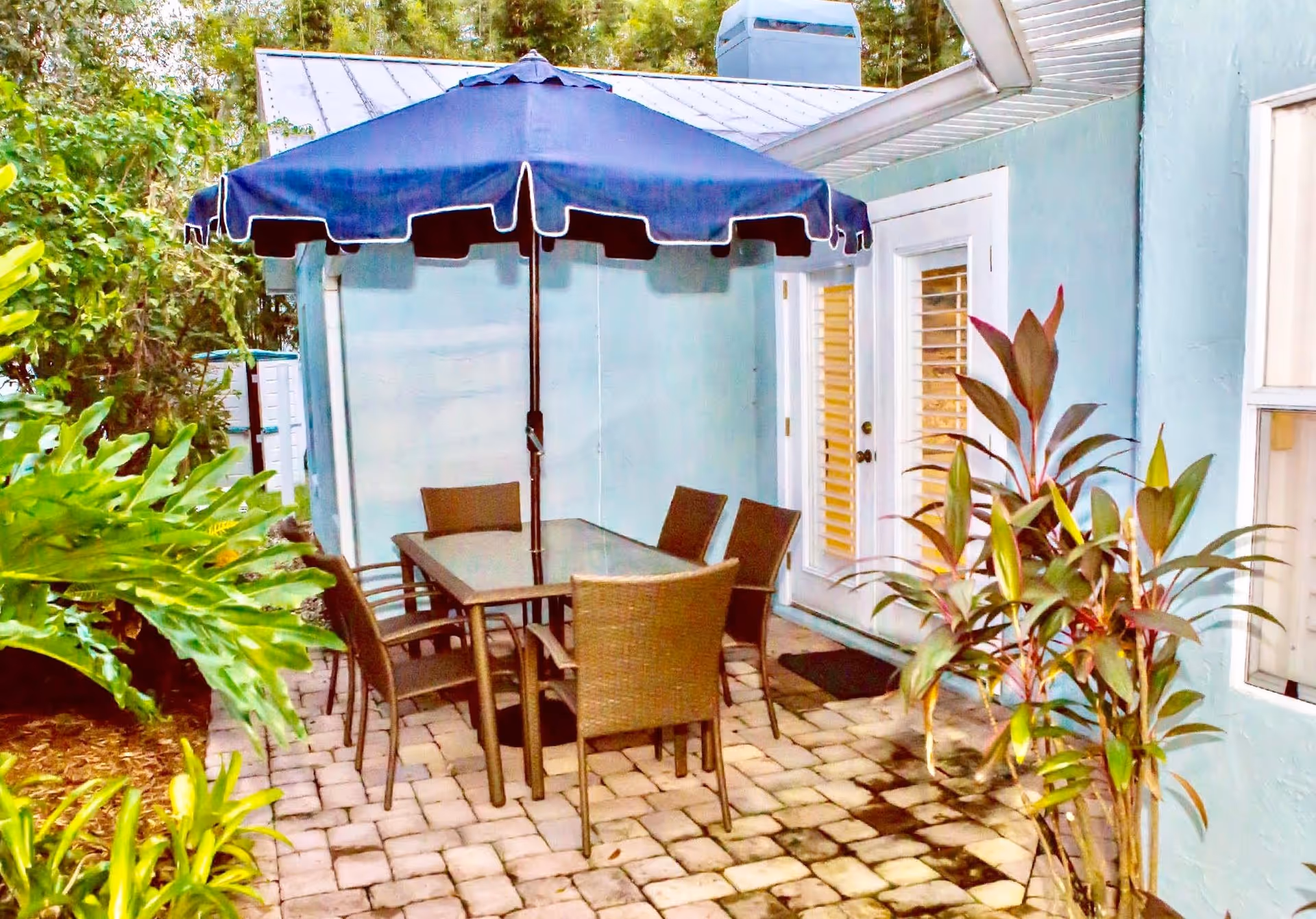 Outdoor patio area with a rectangular glass-top table surrounded by six wicker chairs. A large blue umbrella is positioned over the table. The patio is paved with stone tiles and is adjacent to a light blue building with white-framed windows and a door. There are green plants and foliage around the patio.