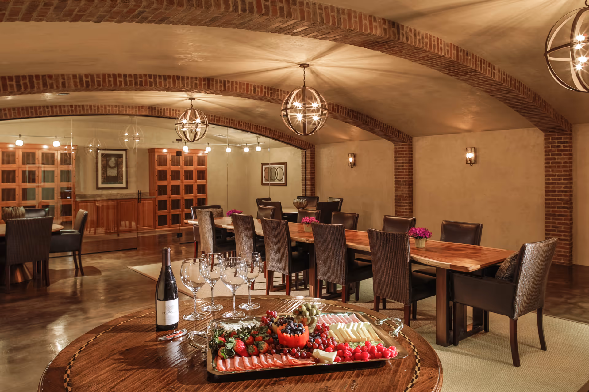 A warmly lit dining room with a long wooden table surrounded by dark chairs. The ceiling features brick arches and modern spherical chandeliers. In the foreground, a round wooden table holds a tray with assorted cheeses, fruits, and meats, along with a bottle of wine and several empty wine glasses. The back wall has wooden cabinets and framed artwork.
