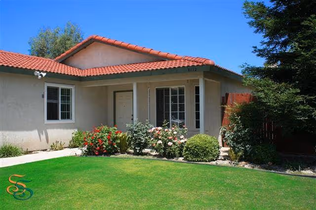 Single-story beige building with a red tile roof, front lawn, and flower beds in front of the entrance.