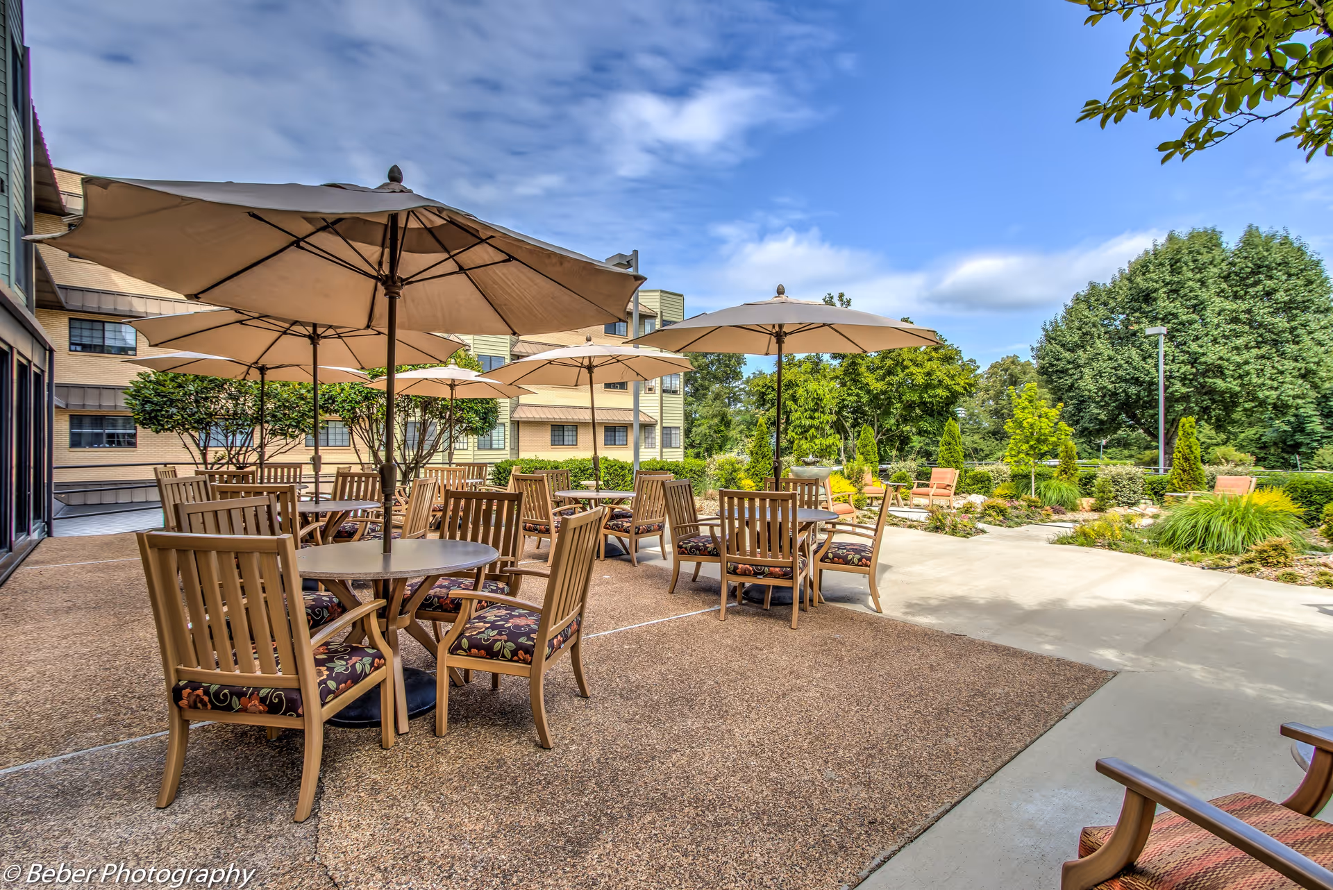 Outdoor patio with wooden tables and chairs under large umbrellas beside landscaped grounds and an adjacent building.