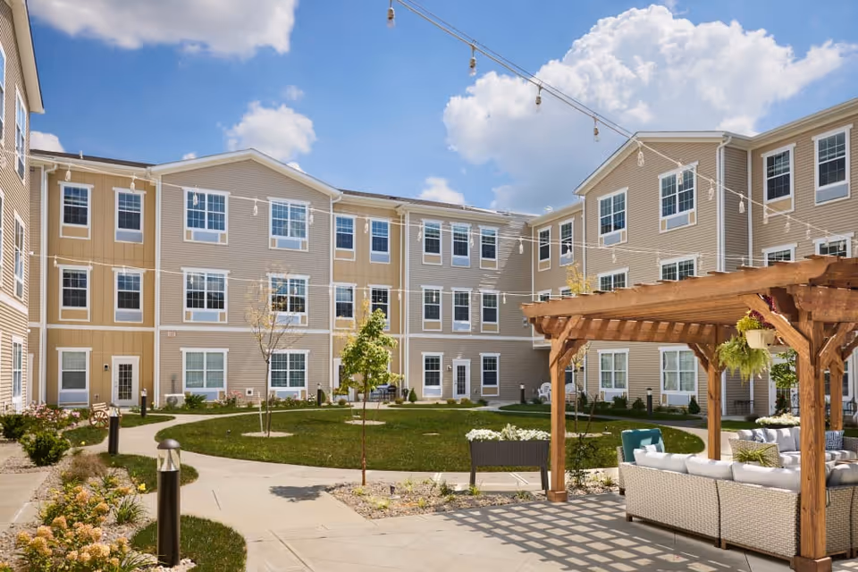 Outdoor courtyard area of a senior living facility with a three-story beige building surrounding a green lawn. There are string lights hanging overhead and a wooden pergola with cushioned seating underneath. Small trees and landscaped flower beds line the paved walkways.