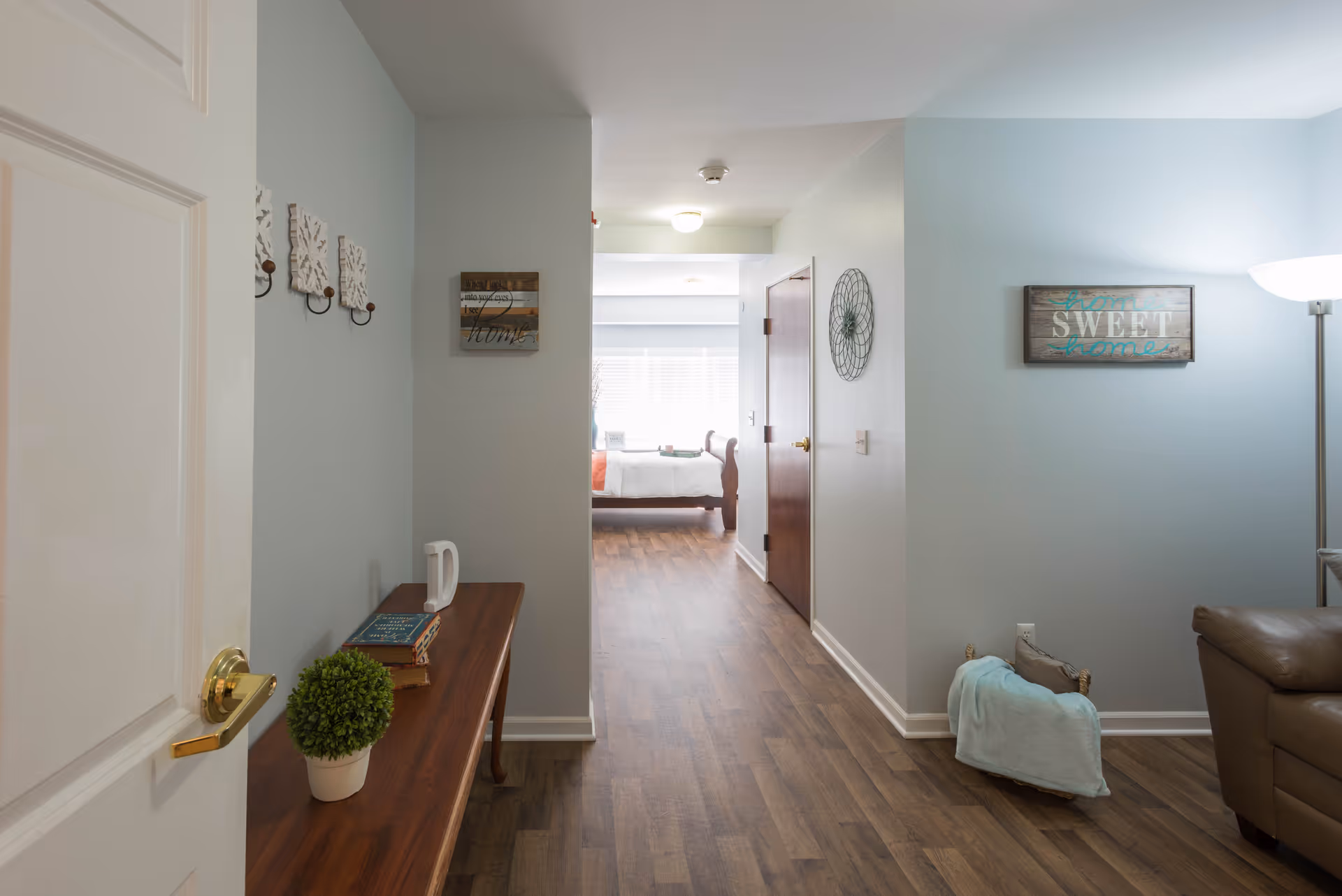 View down a hallway in a senior living facility with light blue walls and wood flooring. On the left, there is a wooden console table with a small potted plant, books, and a decorative letter 'D'. Wall decorations include hooks and framed signs. At the end of the hallway, a bedroom with a bed and window blinds is visible. On the right, part of a brown leather chair and a floor lamp can be seen, along with a sign that reads 'Home Sweet Home'.