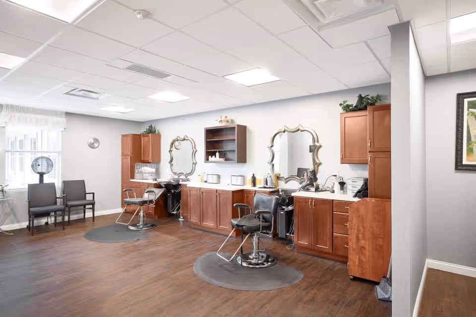 A bright and clean salon area inside a senior living facility with two styling chairs in front of large decorative mirrors, wooden cabinets along the wall, and two waiting chairs near a window with sheer curtains. The floor is wood, and the ceiling has recessed lighting.