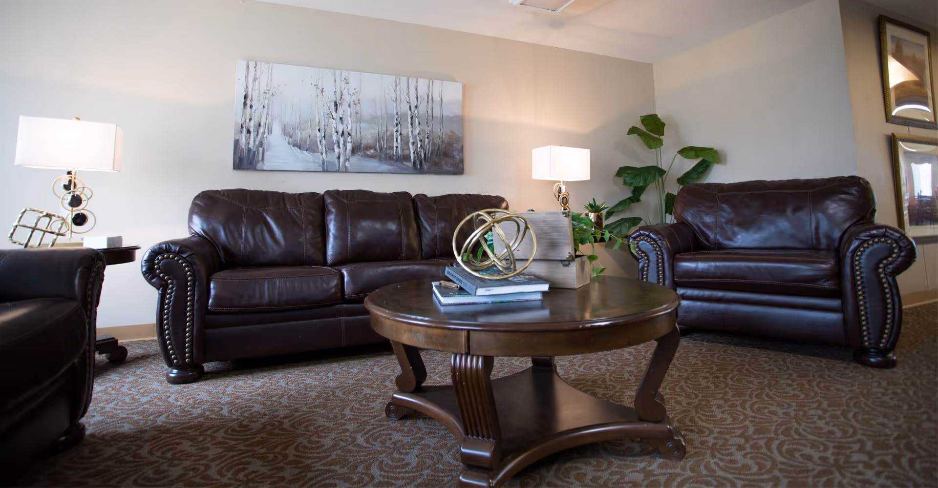 A living room with dark leather sofas and armchairs arranged around a wooden coffee table, lit by table lamps and wall art.