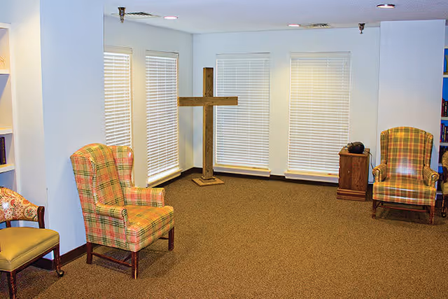 A quiet room with beige carpet and white walls featuring three large windows with closed blinds. There are two plaid upholstered armchairs and a wooden cross standing in the corner. A small wooden cabinet with a telephone on top is also visible.