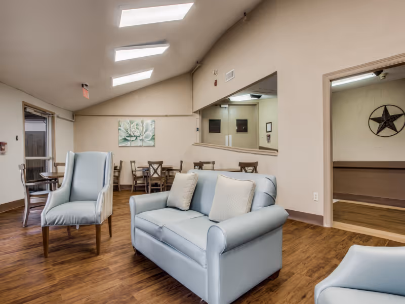 A well-lit common area in a senior living facility featuring light blue cushioned seating including a loveseat and armchairs, wooden flooring, several wooden tables and chairs in the background, a large mirror on one wall, and a decorative painting of a flower. There is also a doorway leading to another room with a star decoration on the wall.