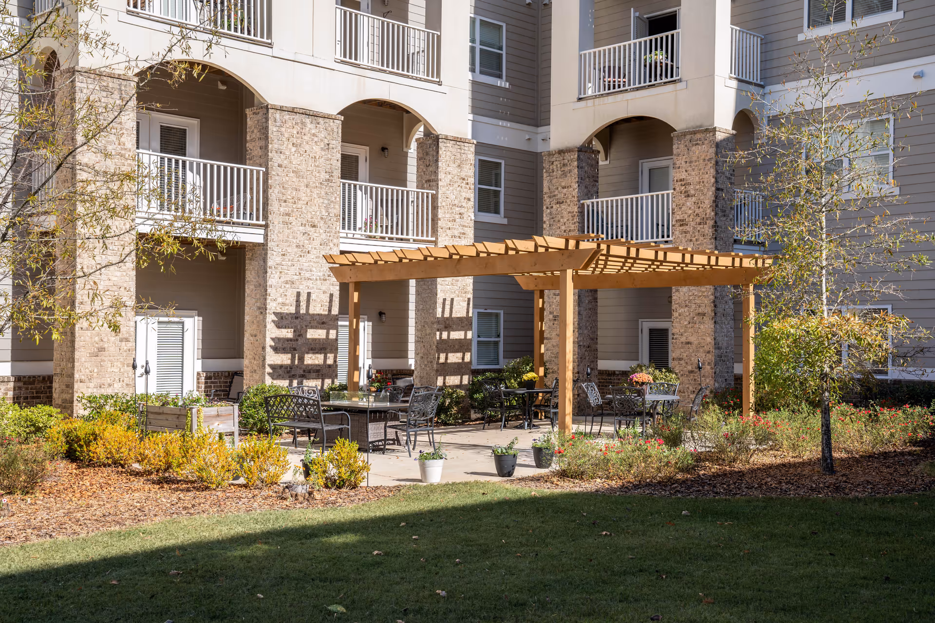 Courtyard with a wooden pergola, outdoor tables and chairs, potted plants, and the exterior of a multi-story residential building with balconies.