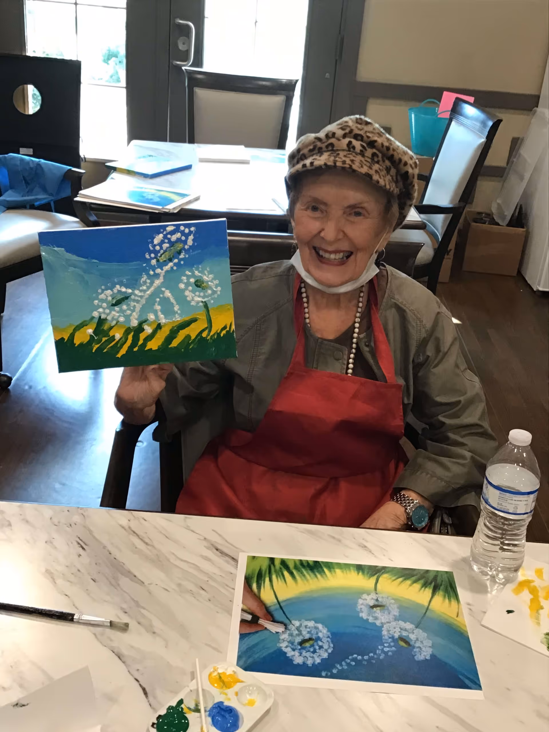 An elderly woman wearing a leopard print hat and red apron is sitting at a table holding up a painting of white flowers against a blue and green background. She is smiling and there are painting supplies, a water bottle, and another painting on the table in front of her. The room has chairs and tables with natural light coming through the windows.