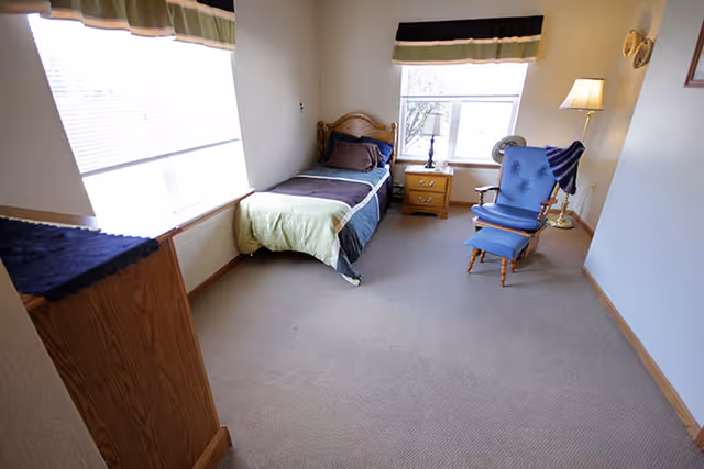 Small bedroom with a single bed against the wall, a nightstand between two windows, and a blue upholstered chair with ottoman.