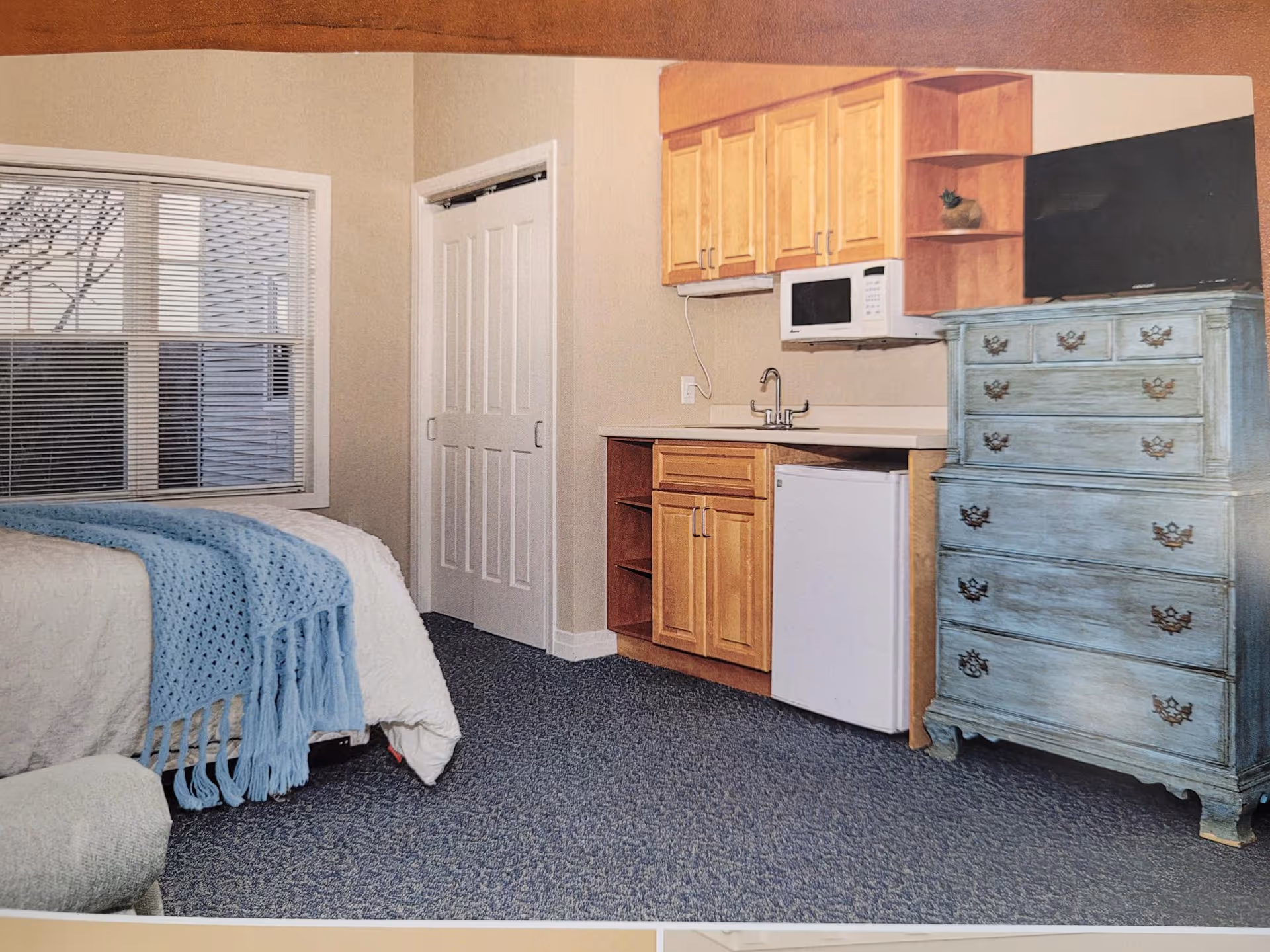 Small studio bedroom with a bed by a window, a kitchenette (sink, microwave, mini-fridge), and a blue distressed dresser.