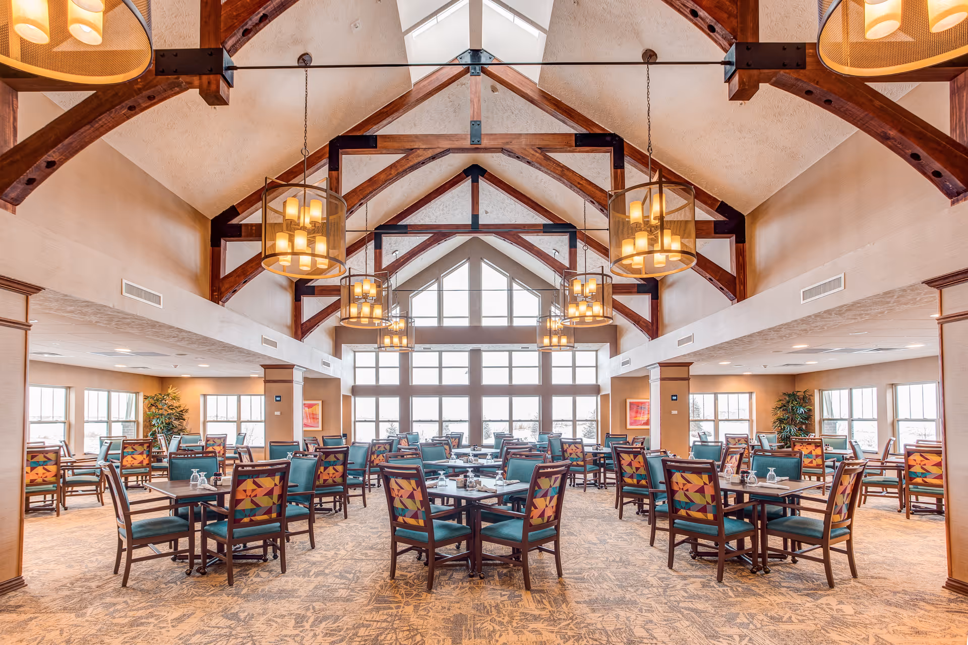 Spacious dining room with multiple tables and chairs featuring colorful patterned backs and teal seats. The room has a high vaulted ceiling with exposed wooden beams and large hanging light fixtures. Large windows at the far end allow natural light to fill the space, and there are plants and framed artwork on the walls.