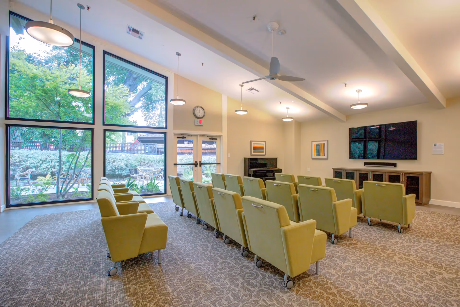 A spacious common area with rows of green cushioned chairs on wheels facing a large flat-screen TV mounted on the wall. The room has large floor-to-ceiling windows letting in natural light and offering a view of an outdoor garden area with trees and patio furniture. The ceiling features multiple hanging light fixtures and a ceiling fan. There is a piano against one wall and framed artwork on the walls.