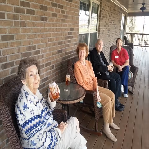 Four people sitting on wicker chairs on a covered wooden porch. Three elderly individuals are seated together, two women and one man, each holding a glass of iced tea. A younger woman in a red shirt and blue pants, possibly a staff member, is sitting with them and smiling. The porch has a brick wall on one side and a railing on the other, with ceiling fans overhead.