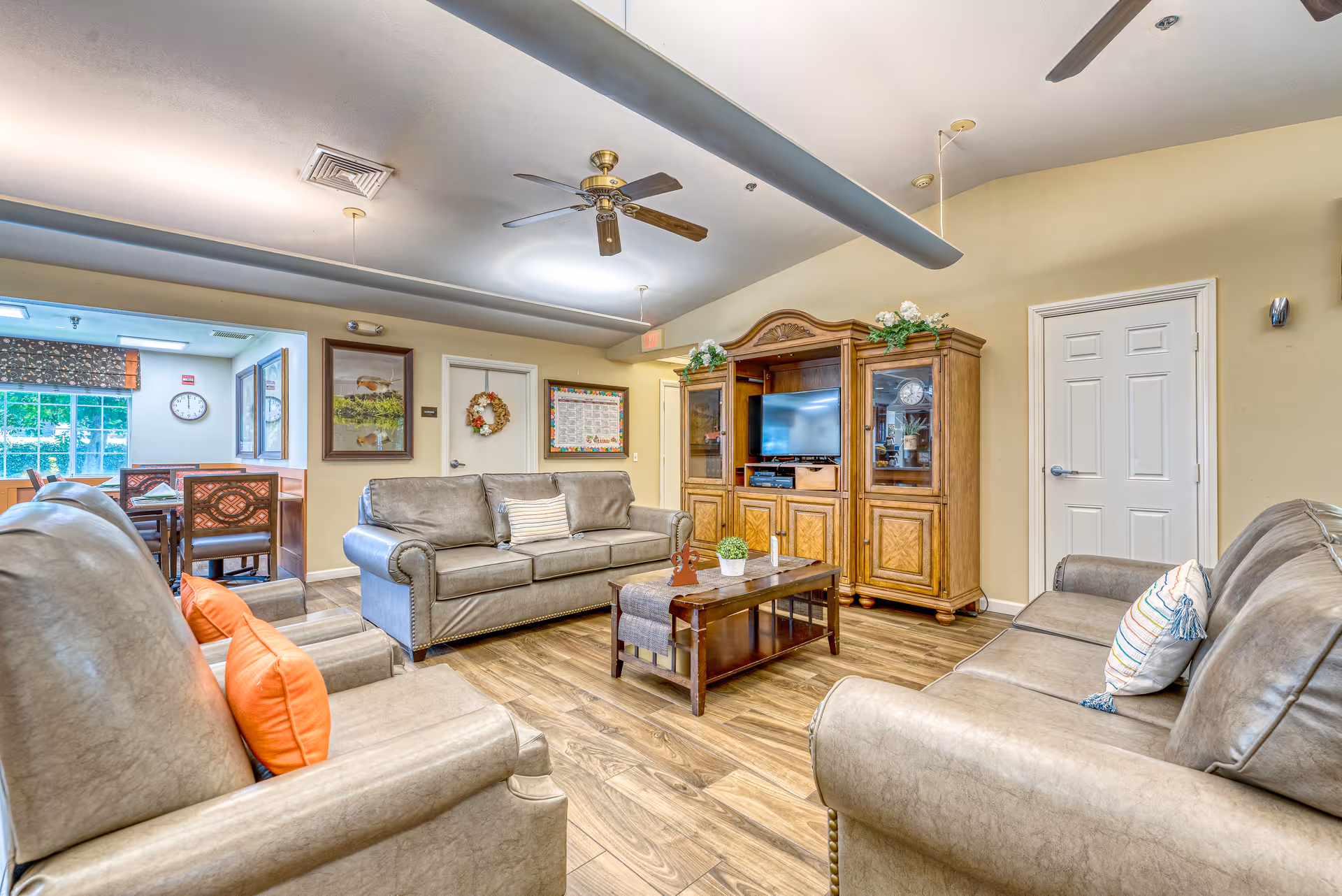 A bright communal living room with sofas arranged around a coffee table facing a wooden entertainment center with a TV.