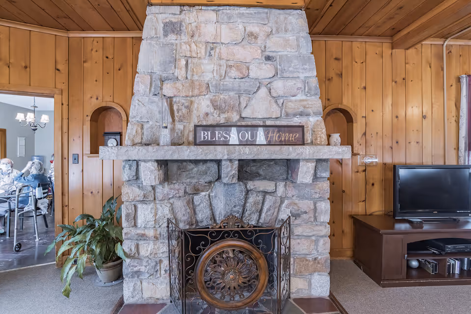 A cozy living room with a large stone fireplace in the center. On the mantel above the fireplace is a sign that reads 'BLESS OUR Home'. To the right of the fireplace is a wooden TV stand with a flat-screen television and some electronic devices. The walls and ceiling are covered in wood paneling. To the left, through an open doorway, a dining area is visible with elderly people seated at a table.