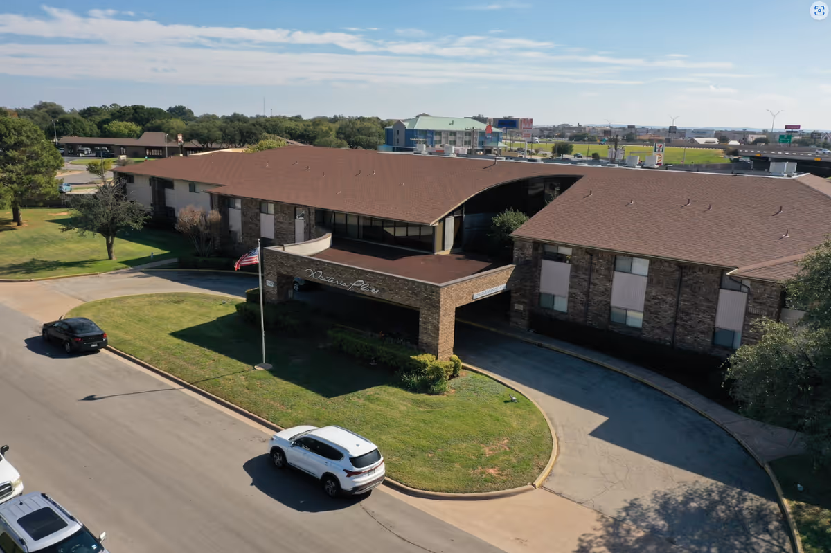 Aerial view of Wisteria Place Independent Living facility showing a two-story brick building with a brown roof, a covered entrance with the facility name displayed, a curved driveway, parked cars, and surrounding greenery under a partly cloudy sky.