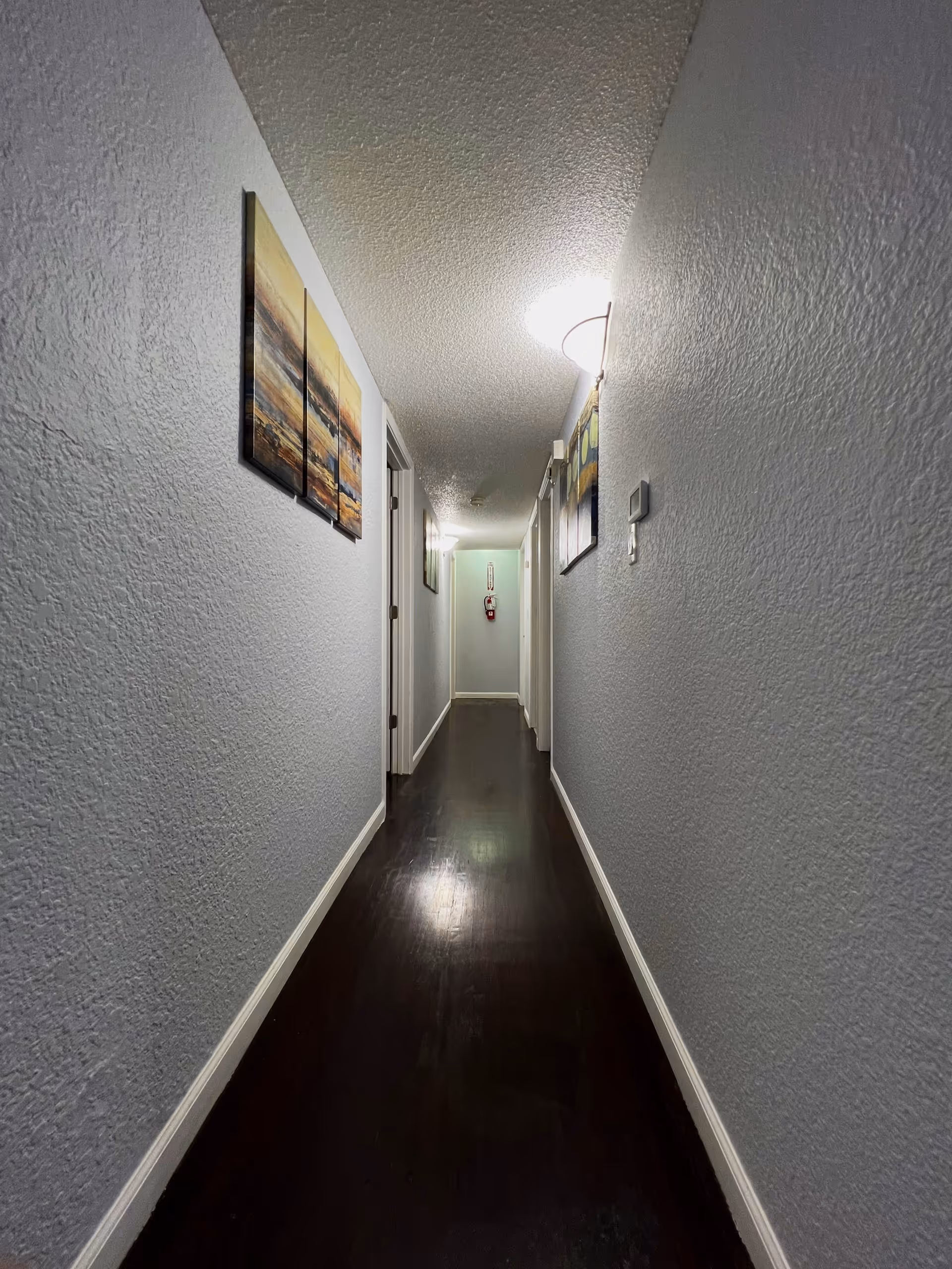 A narrow hallway with dark wooden flooring and light gray textured walls. Several doors line the hallway on both sides, and there are framed artworks hanging on the walls. The hallway is lit by ceiling lights, and a fire extinguisher is mounted on the far wall.