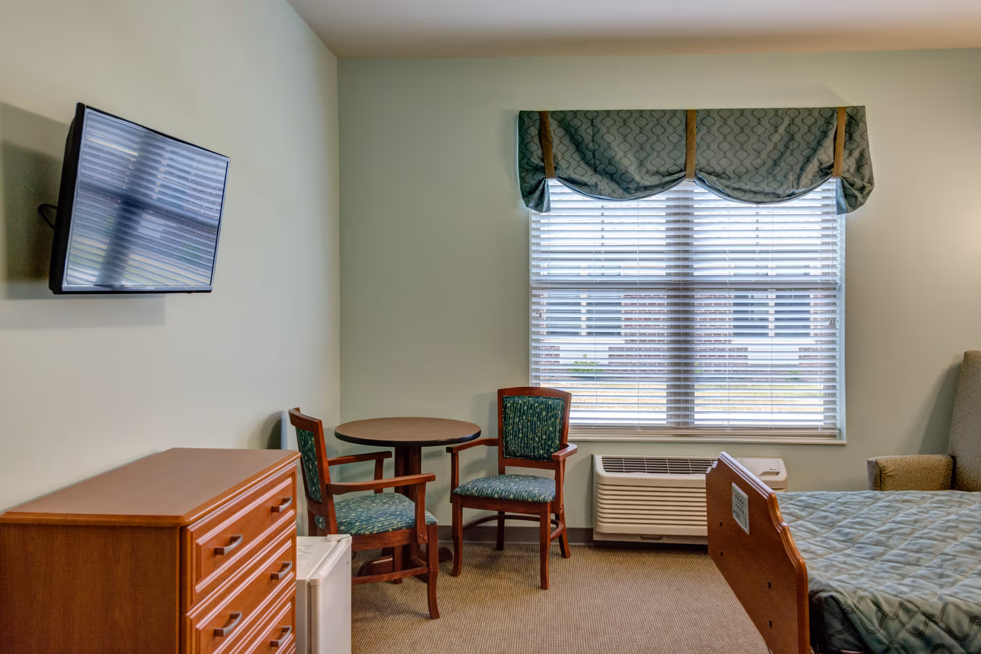 A nursing facility room with a hospital bed partially visible on the right, a window with blinds and a valance, two wooden chairs with green patterned cushions around a small round table, a wooden dresser, a small refrigerator, a wall-mounted flat screen TV, and an air conditioning unit below the window.
