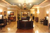A well-lit reception lobby with a central wooden front desk, chandelier, seating areas, and decorative lamps and artwork.