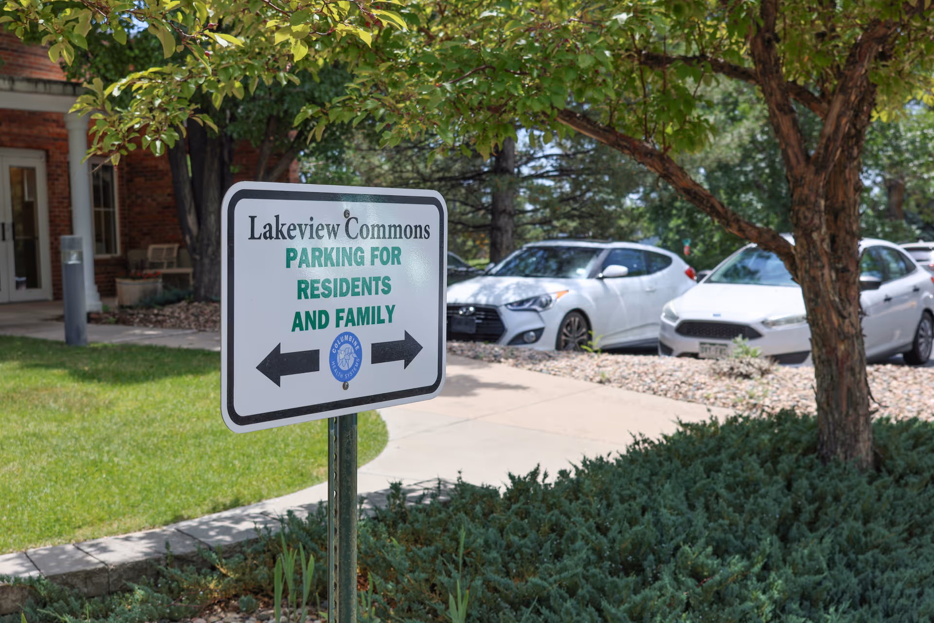 A Lakeview Commons parking sign by a landscaped entrance with parked cars and a tree in front of the building.