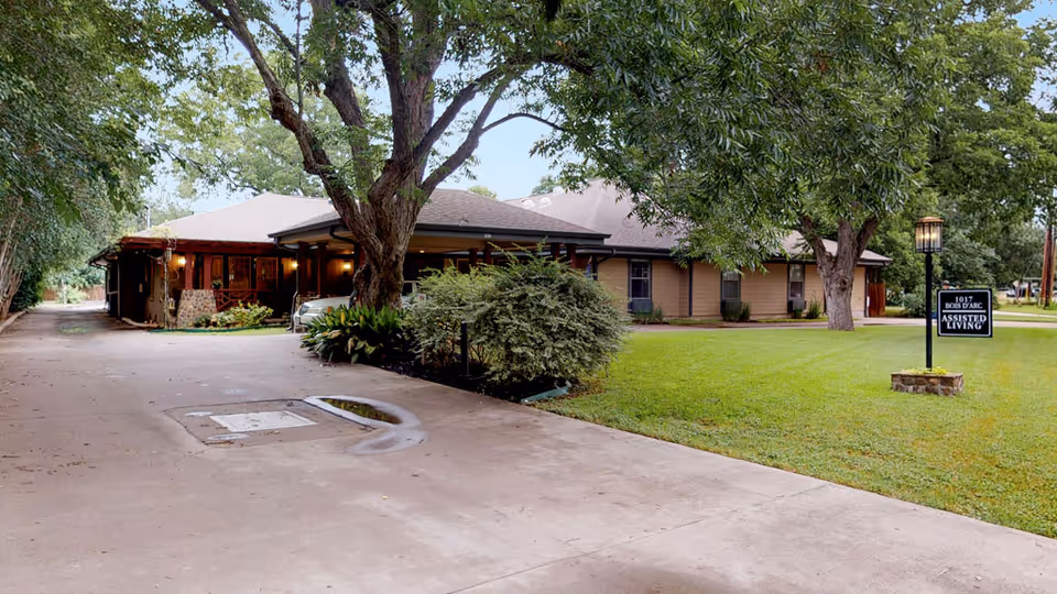 Exterior view of a single-story assisted living facility named Assisted Living Lockhart 1017 Bois D'Arc, surrounded by green grass and large trees, with a paved driveway leading to the building and a sign near a lamp post displaying the facility name.